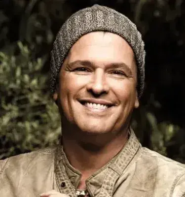 Smiling man wearing a wool cap and beige jacket in a natural environment.