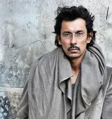 Haider Ackermann con abrigo gris frente a pared envejecida