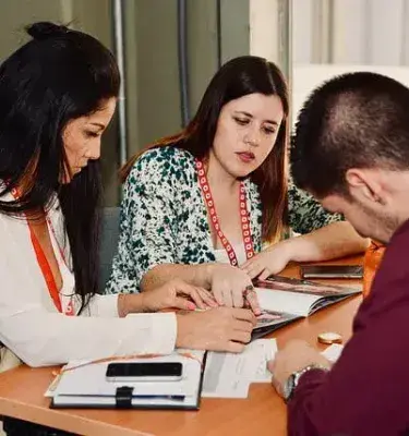 Dos mujeres y un hombre sentados en una mesa, mirando documentos y un folleto.