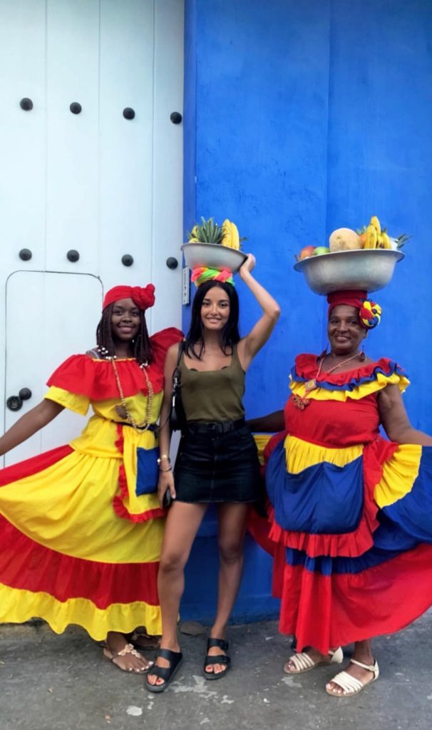Amina Muaddi posando junto dos mujeres palenqueras en Cartagena frente a una casa azul con puerta blanca, moda Colombia, Cartagena 
