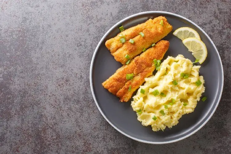Dos filetes de pescado rebozado con puré de papas y limón en un plato gris.