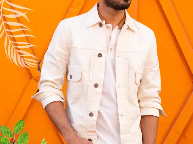 Hombre con chaqueta blanca y camisa, posando frente a una pared naranja.