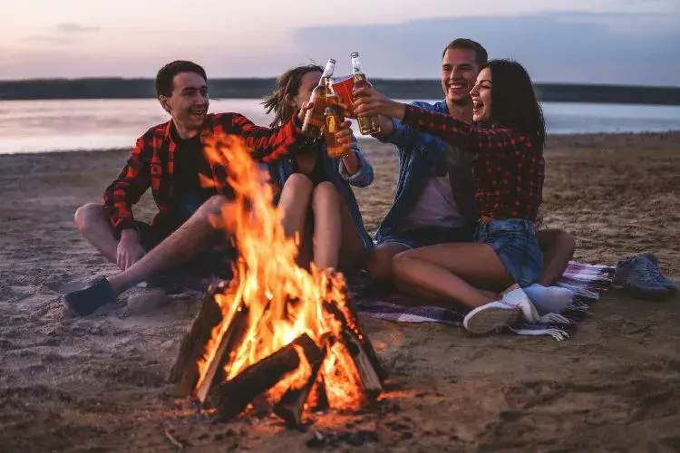 Grupo de amigos brindando con cervezas alrededor de una fogata en la playa al atardecer